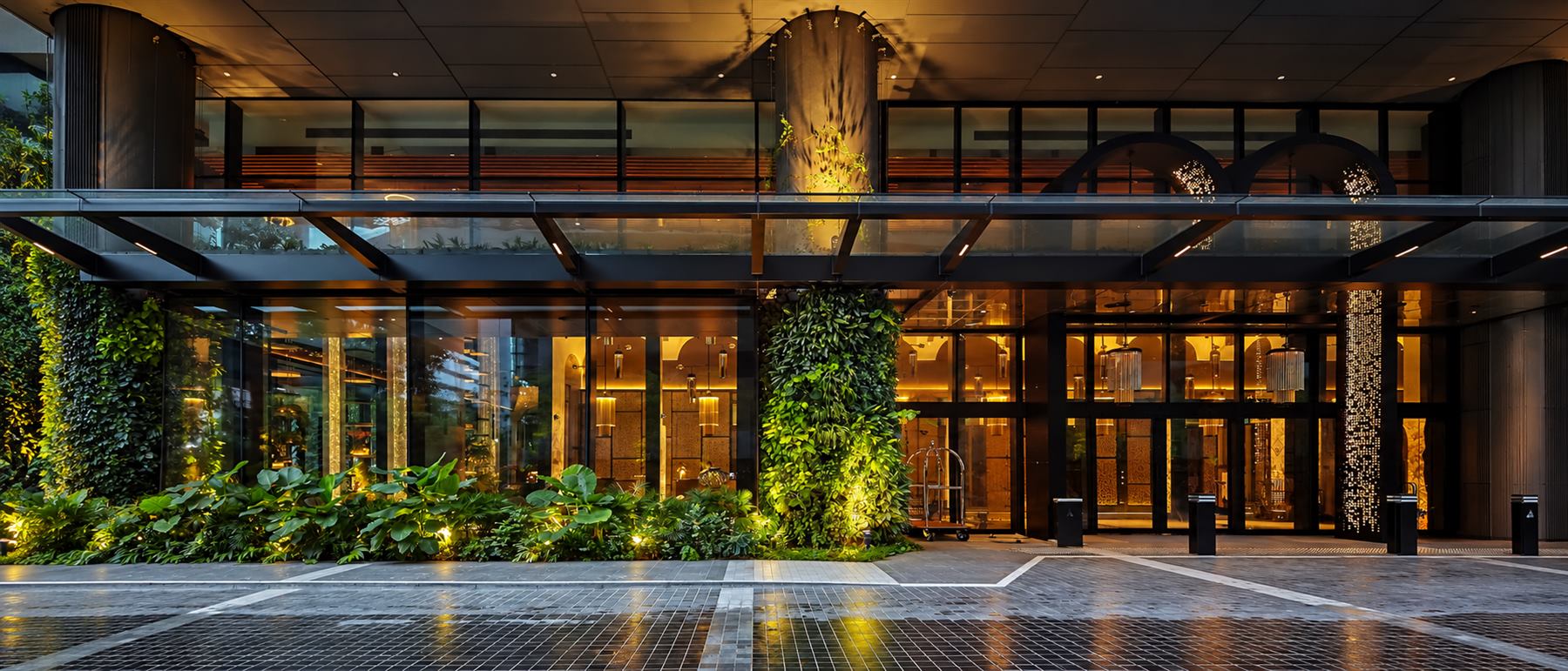 Artyzen Singapore arrival court at dusk — glass porte-cochère, planted green walls, warmly lit lobby with fluted glass pendants and perforated screens beyond