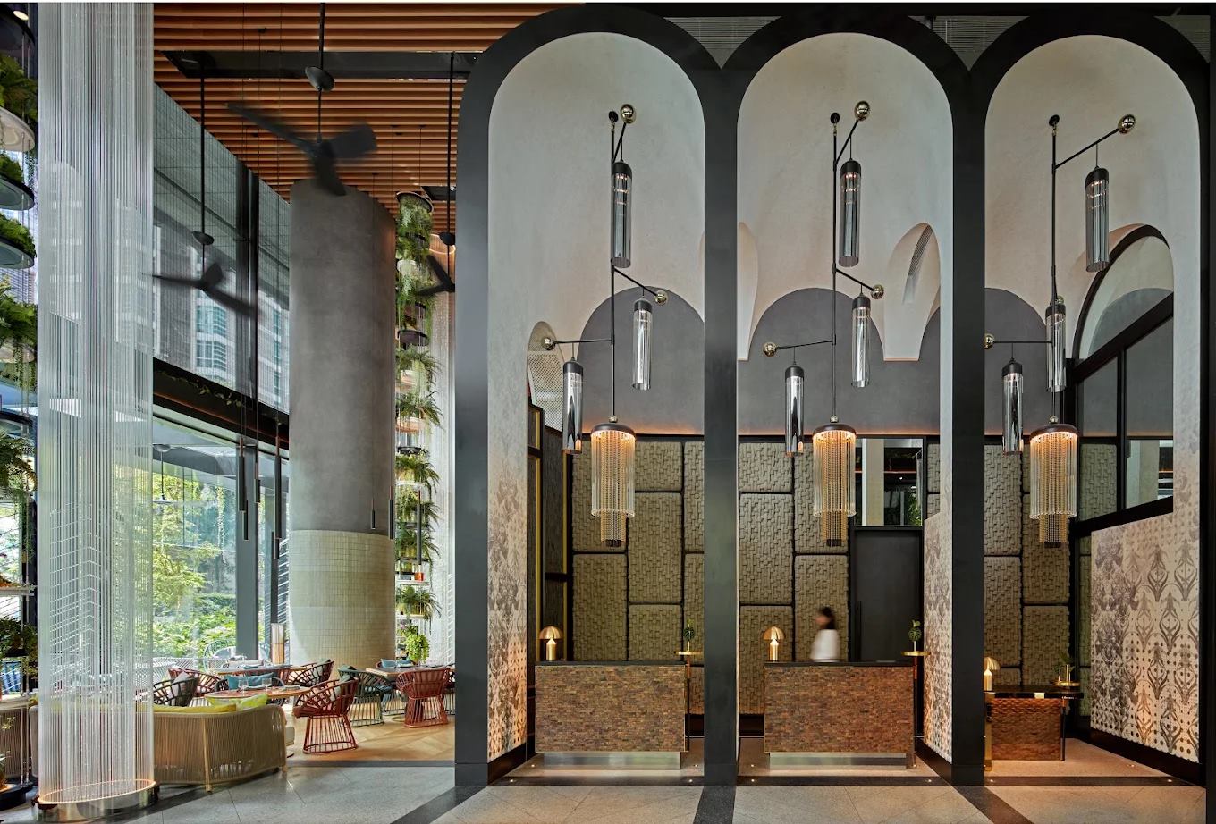 Hotel lobby with tall dark arches, fluted glass pendants, woven wall panels and a planted double-height atrium beyond