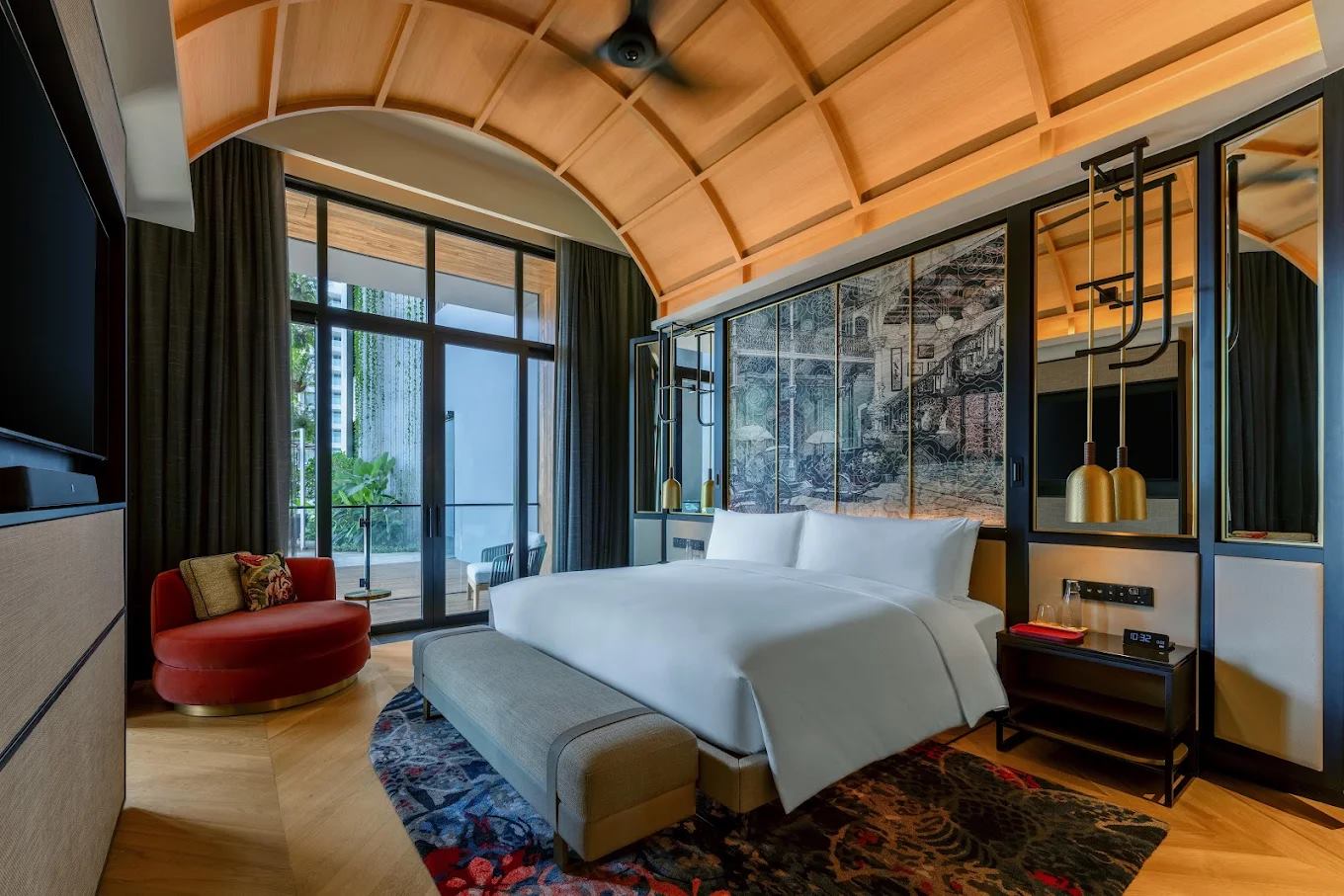 Hotel guest room with a barrel-vaulted timber ceiling, etched screen behind the bed, brass pendants, red velvet chair and a patterned rug
